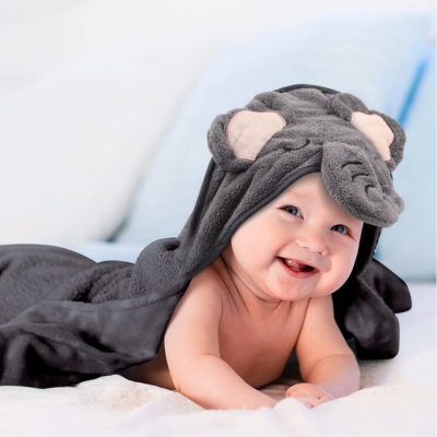 Un bébé souriant allongé sur une surface moelleuse, enveloppé dans une cape de bain bébé en forme d'éléphant gris. La cape de bain bébé est dotée d'une capuche avec des oreilles d'éléphant et une trompe en relief, ajoutant une touche ludique et adorable.