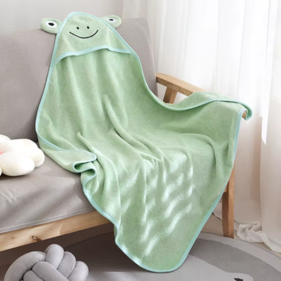 Une cape de bain bebe verte avec une capuche en forme de grenouille souriante est posée sur un canapé gris clair, dans une pièce lumineuse au décor doux et épuré.