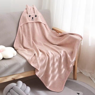 Une cape de bain bebe en tissu rose pâle, décorée d'une capuche avec des oreilles de lapin et un visage brodé, est étalée sur un canapé.