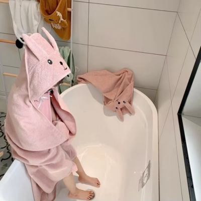 Une petite fille, assisse sur le bord de la baignoire, enveloppée dans sa cape de bain bebe rose pâle avec la capuche de lapin sur sa tête.