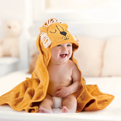 Un enfant souriant est assis portant une cape de bain bebe de couleur orange. Sa tête est coiffée d'une capuche en forme de lion avec des franges.