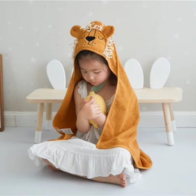 Une petite fille porte une cape de bain bebe orange en forme de lion, avec une capuche à crinière. Elle est assise, tenant un jouet à la main.