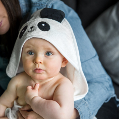 Un bébé enveloppé dans une cape de bain bebe blanche en coton doux, avec une capuche en forme de tête de panda et des oreilles noires, assis confortablement sur les genoux de sa maman. Un moment de tendresse après le bain, mettant en avant le confort et la douceur de la cape.