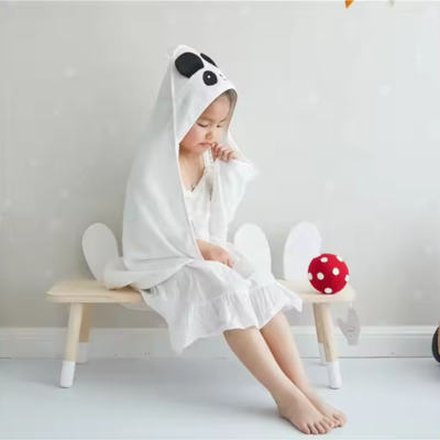 Une fillette paisiblement assise sur un banc, drapée dans une cape de bain bebe blanche avec une capuche panda. Elle semble paisible et détendue, illustrant la taille généreuse et le confort de la cape.